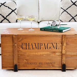 Könnte beinhalten: Eine Holzkiste mit Metallverzierungen. Die Kiste ist braun lackiert und hat die Aufschrift "Champagne, Produit de France, 1937" auf der Vorderseite. Die Kiste steht auf einem weißen Teppich vor einem weißen Sofa.