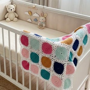 May include: A white crib with a colorful, crocheted blanket draped over the side. The blanket features a pattern of large, colorful circles in pink, teal, navy, and gold. A crocheted teddy bear and a bunny rattle are visible.