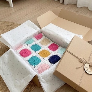 May include: A colorful, handmade crochet blanket with a polka dot design, packaged in a cardboard box with tissue paper. The blanket features pink, teal, yellow, and navy blue circles. The box is tied with twine.