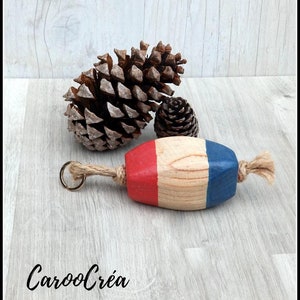 Op de afbeelding: Een houten nautische boeien sleutelhanger met rode, witte en blauwe verf. De boei is bevestigd aan een touw en een gouden sleutelring. De sleutelhanger ligt op een houten oppervlak met dennenappels.