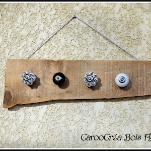 Op de afbeelding: Een houten bord met vier decoratieve knoppen, twee wit en twee zwart, met een bloemmotief. Het bord hangt aan een wit touw en heeft de tekst "CarooCréa Bois Flotte" onderaan.
