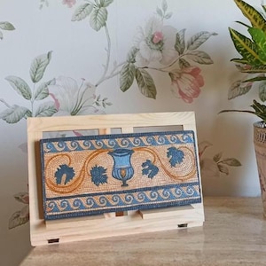 Può includere: Un espositore in legno con una pochette blu e oro con motivo. La pochette ha un design con un vaso con viti e foglie.