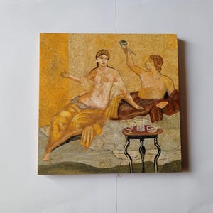 Pode incluir: Uma obra de arte quadrada e emoldurada que retrata uma cena clássica. Duas figuras são mostradas reclinadas, com uma delas segurando um recipiente. Uma pequena mesa com copos está à frente delas. A obra de arte tem uma paleta de cores quentes e terrosas.