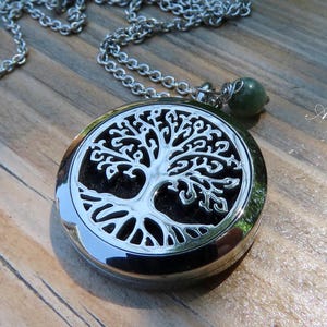 Peut inclure: Collier argenté avec un pendentif rond représentant un arbre de vie. Le pendentif a un intérieur noir et une chaîne argentée. Une petite pierre verte est suspendue près du fermoir. Le collier est posé sur une surface en bois.