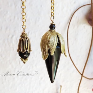 Könnte beinhalten: Zwei schwarze Edelsteinpendel mit goldenen Ketten und dekorativen Akzenten. Die Pendel hängen vor einem weißen Hintergrund mit einer grünen Pflanze im Hintergrund.