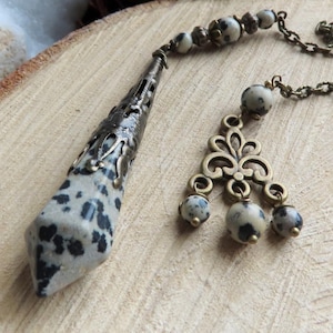 Op de afbeelding: Een pendel met een gespikkelde steen en sierlijk metaalwerk. De pendel heeft een ketting en een decoratief element met kleine, bijpassende stenen. Het ontwerp bevat de tekst "Alexia Creations" op een houten oppervlak.