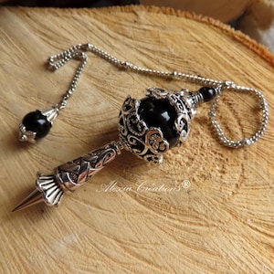 Op de afbeelding: Een zwarte edelsteenpendule met een zilveren ketting en sierlijke zilveren accenten. De pendule hangt op een houten oppervlak.