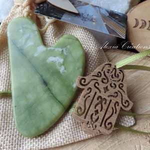Könnte beinhalten: Eine Sammlung natürlicher Wellness-Artikel. Enthält ein grünes Jade-Gua-Sha-Werkzeug, einen Holzanhänger in Hamsa-Handform und eine Mondphasen-Schnitzerei aus Holz. Die Artikel werden auf einer Holzoberfläche mit einem Jutesack präsentiert.