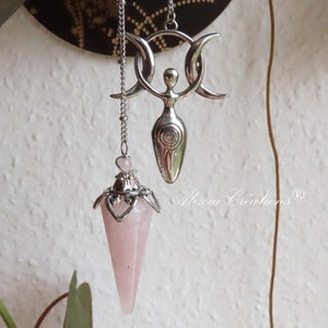 May include: Two silver pendulums with chains. One pendulum features a pink crystal point and the other features a silver triple moon goddess charm. The charm has a spiral design and the text "Moon Creations" is visible in the image.