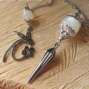 May include: A white crystal pendulum with a silver chain and a silver charm with a leaf design. The pendulum is on a wooden surface.