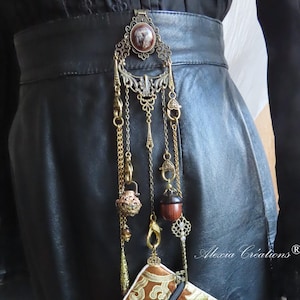 Puede incluir: Accesorio de cinturón decorativo con una pequeña bolsa con estampado marrón y dorado. El accesorio presenta elementos metálicos dorados, cadenas y abalorios, incluyendo una bellota y un colgante decorativo. La bolsa tiene una cremallera negra y una borla roja.