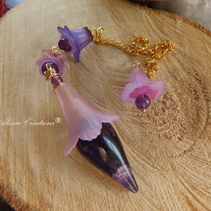 Può includere: Un pendolo fatto a mano con un design floreale viola e rosa. Presenta una catena dorata e perline di ametista. Il corpo principale è a forma di goccia viola scuro, racchiuso in un involucro rosa a forma di fiore.