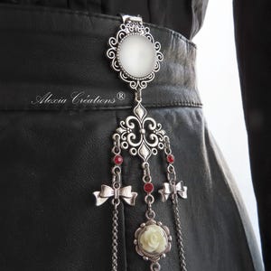 May include: A silver-toned belt clip with a white oval center and ornate filigree design. It features dangling chains with red beads, small bows, and a white rose pendant. The text "Alexia Creations" is visible.