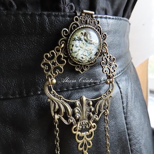 Peut inclure: Boucle de ceinture ornée de couleur bronze avec une pièce centrale circulaire et à motifs. La boucle présente des détails de filigrane et de chaîne complexes, attachés à une ceinture en cuir noir. Le texte "Alexia Creations" est visible.