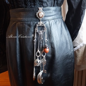May include: A silver-toned belt accessory with dangling charms, including a key, acorn, and moon. The top piece is an ornate pendant with a circular design. The accessory hangs against a black leather skirt.