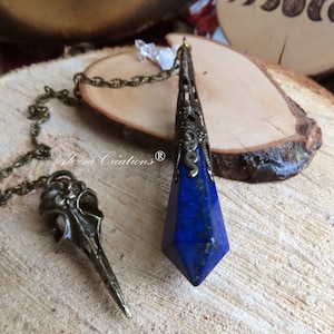 May include: A blue lapis lazuli crystal pendant on a bronze chain with a bird skull charm. The pendant is in the shape of a point and has intricate bronze detailing. The chain is a delicate, linked chain. The bird skull charm is detailed and has a pointed beak.