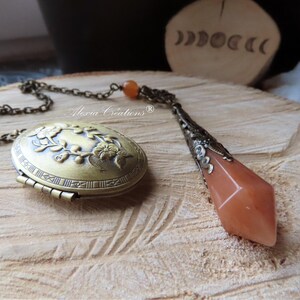 May include: A bronze chain necklace with a locket and a carnelian crystal pendulum. The locket is oval shaped and features an engraved floral design. The pendulum is faceted and has a bronze setting.