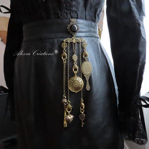May include: A black leather belt with a decorative gold-colored pendant featuring hanging charms, including a pocket watch, a small mirror, and keys. The belt is worn over a black outfit.