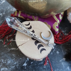 Può includere: Un pendolo in argento e cristallo con un ciondolo a forma di mezzaluna appeso a una catena. Il pendolo è appoggiato su una fetta di legno con le parole "Alexu Creations" e tre mezze lune intagliate.