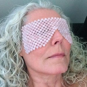 Peut inclure: Un masque pour les yeux rose clair composé de petites perles rondes. Le masque couvre les yeux et une partie du nez. Le masque est porté par une personne aux cheveux bouclés gris. La marque "Alexia Créations" est visible en bas.