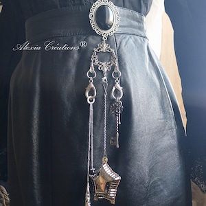 May include: A black leather belt with a silver-toned brooch featuring a black oval stone. Dangling from the brooch are chains with charms, including a star, a key, and a teardrop. The text "Alexia Créations" is visible.