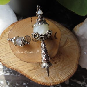 Peut inclure: Un ensemble de pendule et de boucles d'oreilles assorties de couleur argentée, avec une pierre arrondie nacrée. Le pendule a une longue poignée ornée et une pointe. La boucle d'oreille est une version plus petite du pendule.