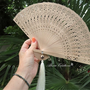 May include: A light brown, wooden hand fan with intricate cut-out designs of moons and stars. The fan is held open, revealing its delicate structure and a white tassel. The fan is a decorative accessory.