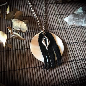 May include: A white and black round pendant necklace with a silver charm and black fringe. The necklace is on a woven bamboo mat.