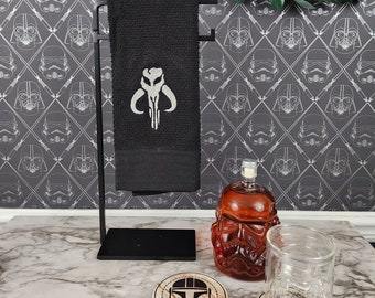 Símbolo del Mythosaur mandaloriano bordado en plata sobre un paño de cocina negro.