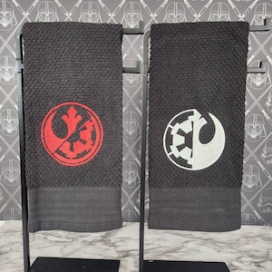 May include: Two black hand towels with embroidered Star Wars symbols, one red and one white. The towels are displayed on black metal stands against a gray Star Wars-themed wallpaper.