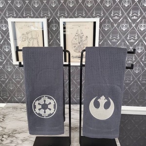 Könnte beinhalten: Zwei graue Küchentücher mit gestickten Star Wars-Logos hängen an einem schwarzen Metallständer. Das Handtuch links hat das imperiale Symbol, das Handtuch rechts das Symbol der Rebellenallianz. Die Handtücher werden vor einer Wand mit einem sich wiederholenden Muster von Darth Vaders Helm ausgestellt.