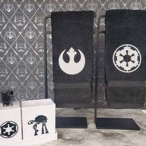 Peut inclure: Serviettes de toilette noires avec des symboles blancs brodés, présentées sur un support en métal noir. Les serviettes présentent les logos de l'Alliance Rebelle et de l'Empire Galactique. Des boîtes blanches avec des graphiques Star Wars sont également visibles.