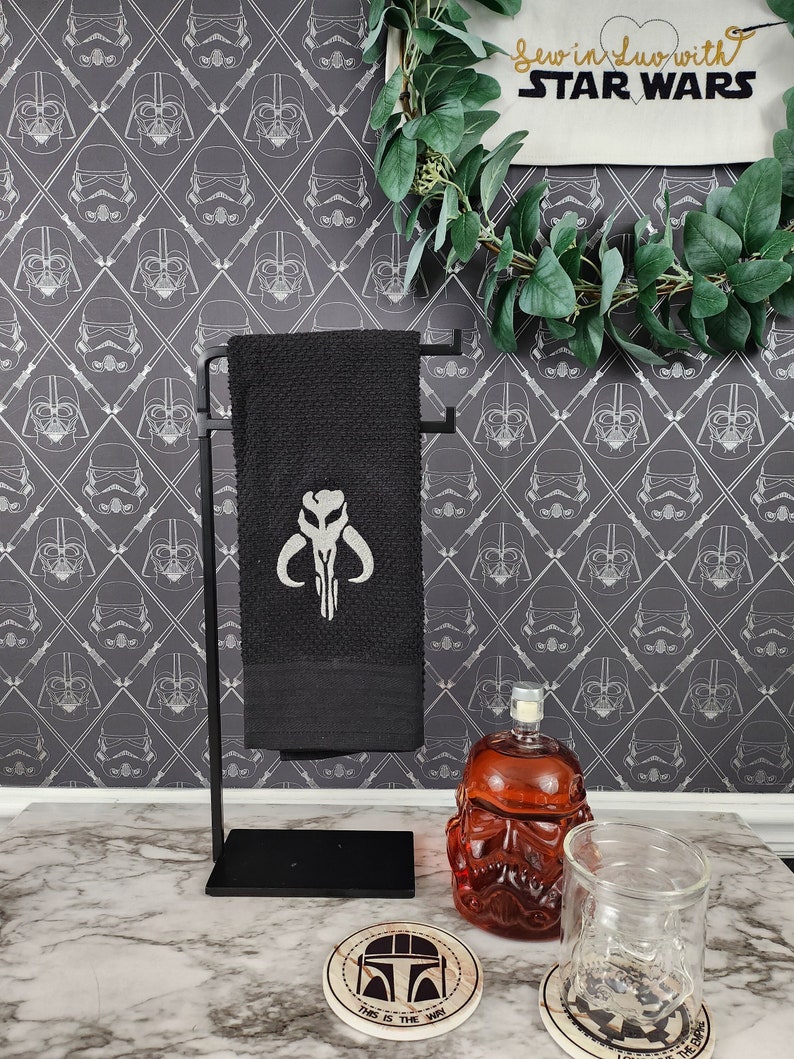 Puede incluir: Una toalla de mano negra con un s&iacute;mbolo mandaloriano blanco se muestra en un toallero de metal negro. El fondo presenta un papel pintado tem&aacute;tico de Star Wars con dise&ntilde;os de Darth Vader y Stormtroopers. Tambi&eacute;n se ven posavasos y una jarra.