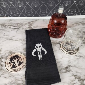 Puede incluir: Una toalla de mano negra bordada con un s&iacute;mbolo de calavera mandaloriana blanca. Tambi&eacute;n hay una jarra roja con forma de calavera, un vaso y dos posavasos con dise&ntilde;os tem&aacute;ticos de Star Wars. Un posavasos dice "THIS IS THE WAY."