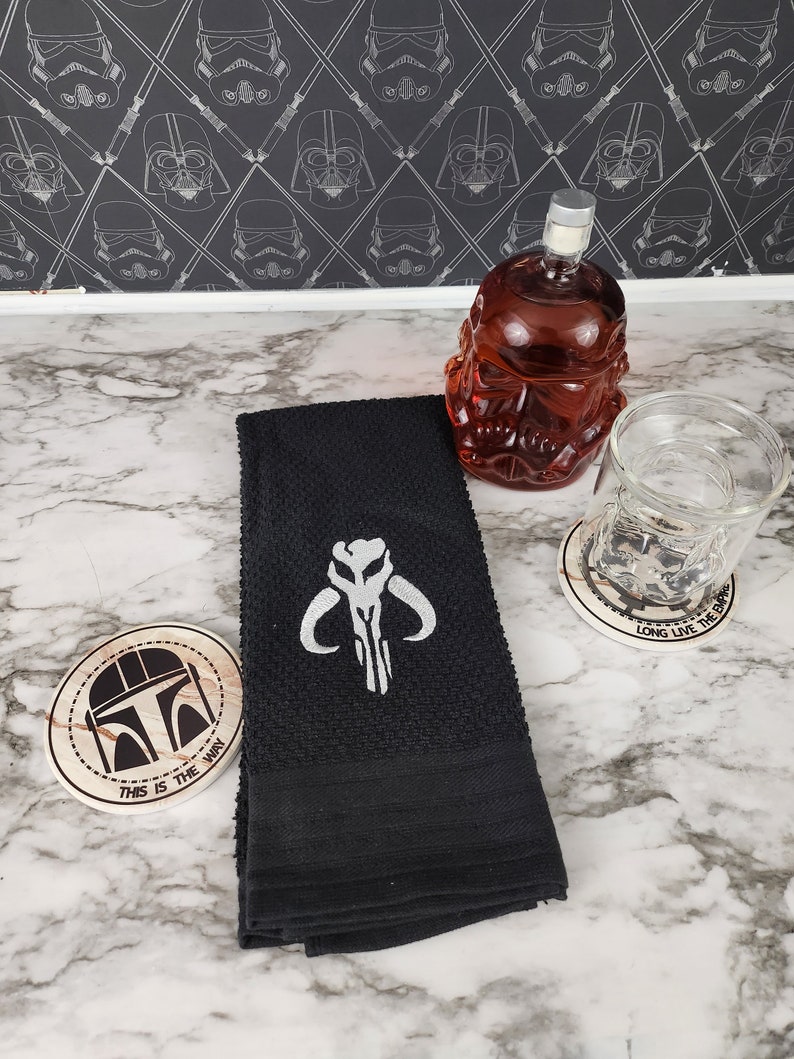 Puede incluir: Una toalla de mano negra bordada con un s&iacute;mbolo de calavera mandaloriana blanca. Tambi&eacute;n hay una jarra roja con forma de calavera, un vaso y dos posavasos con dise&ntilde;os tem&aacute;ticos de Star Wars. Un posavasos dice "THIS IS THE WAY."