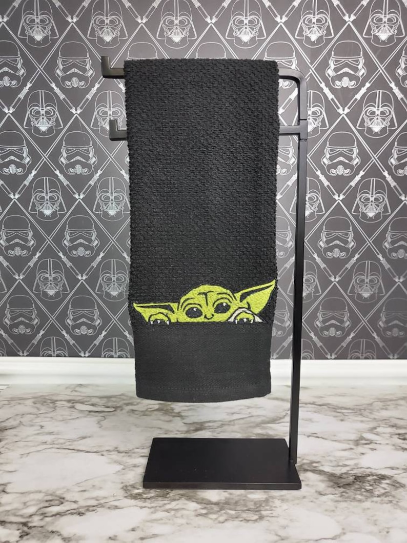 Puede incluir: Toalla de cocina negra con un Baby Yoda verde asomando por detr&aacute;s de la toalla. La toalla est&aacute; colgada en un toallero de metal negro.