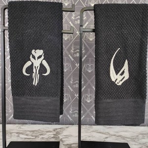 Puede incluir: Dos toallas de mano negras con diseños bordados en blanco, exhibidas en soportes de metal negro. Una toalla presenta un emblema en forma de calavera, la otra un casco estilizado. El fondo muestra un papel pintado temático de Star Wars.
