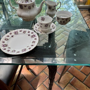 Puede incluir: Un juego de té de porcelana blanca con diseños florales morados. El juego incluye una tetera, una crema, un azucarero, una taza, un platillo y un plato. El juego de té se exhibe sobre una mesa de vidrio.