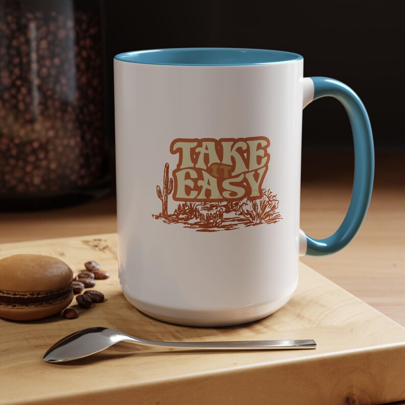 Puede incluir: Taza de cer&aacute;mica blanca con interior y asa azules. La taza presenta el texto "TAKE EASY" en una fuente retro con una ilustraci&oacute;n de paisaje des&eacute;rtico. Un macaron, granos de caf&eacute; y una cuchara est&aacute;n sobre una superficie de madera.