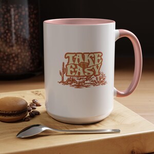Puede incluir: Taza de cer&aacute;mica blanca con interior y asa rosas. La taza presenta el texto "TAKE EASY" en una fuente retro con una ilustraci&oacute;n de paisaje des&eacute;rtico. Un macaron, granos de caf&eacute; y una cuchara est&aacute;n sobre una superficie de madera.