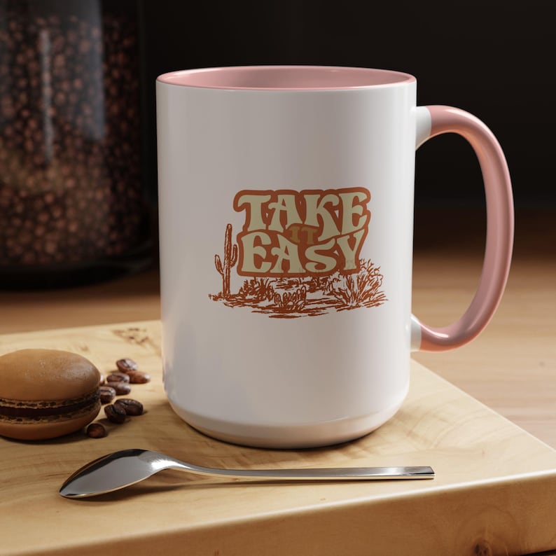 Puede incluir: Taza de cer&aacute;mica blanca con interior y asa rosas. La taza presenta el texto "TAKE EASY" en una fuente retro con una ilustraci&oacute;n de paisaje des&eacute;rtico. Un macaron, granos de caf&eacute; y una cuchara est&aacute;n sobre una superficie de madera.