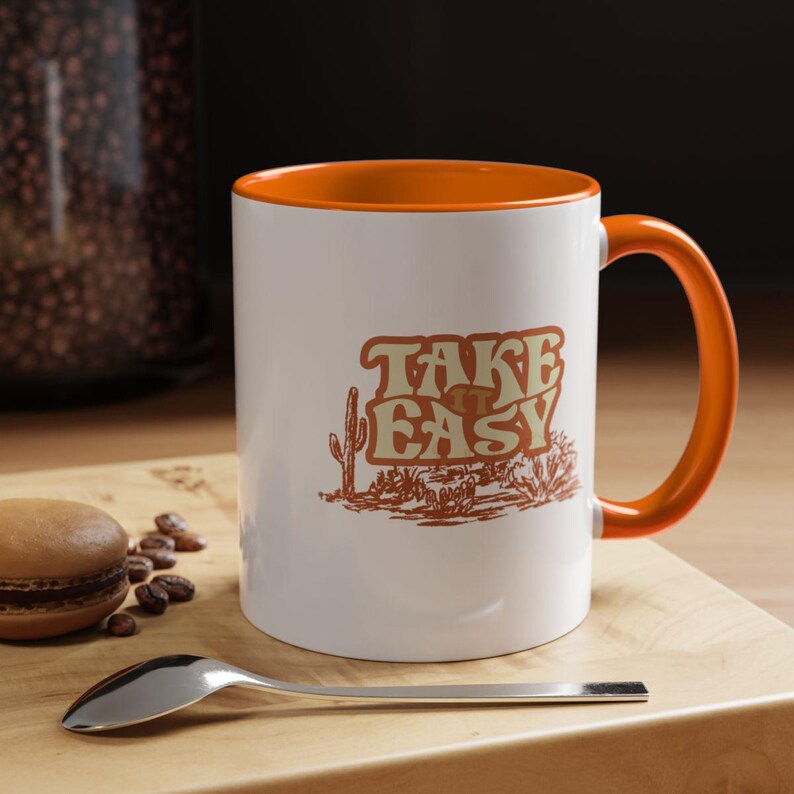 Puede incluir: Taza de cer&aacute;mica blanca con interior y asa naranja. La taza presenta el texto "TAKE EASY" en una fuente retro, junto con una ilustraci&oacute;n de un paisaje des&eacute;rtico en tonos marrones. Una cuchara y un macaron est&aacute;n sobre una superficie de madera.