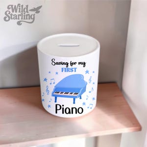May include: A white ceramic cylindrical money box featuring a blue grand piano graphic, musical notes, and stars. The text reads Saving for my FIRST Piano in black and blue lettering. A coin slot is located on the top surface of the container.