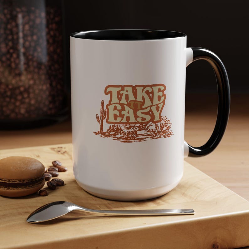 Puede incluir: Taza de cer&aacute;mica blanca con asa e interior negros. La taza presenta el texto "TAKE EASY" en una fuente retro con una ilustraci&oacute;n de paisaje des&eacute;rtico en tonos marrones y beige.
