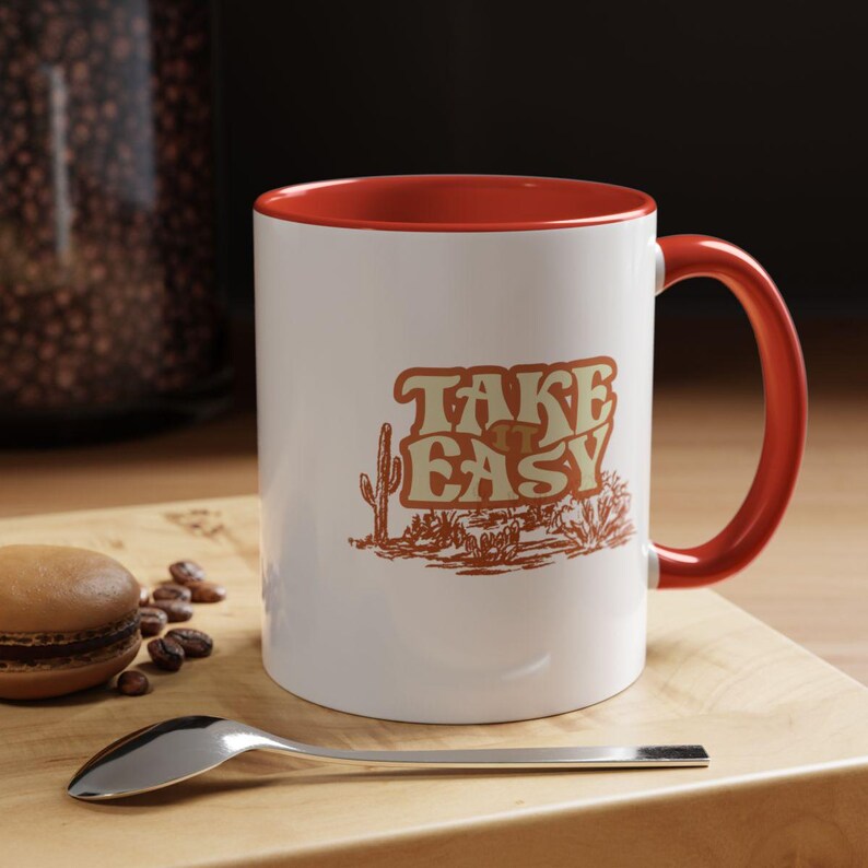 Puede incluir: Taza de cer&aacute;mica blanca con interior y asa rojos. La taza presenta el texto "TAKE EASY" en una fuente retro, junto con una ilustraci&oacute;n de paisaje des&eacute;rtico en tonos marrones. Una cuchara y un macaron est&aacute;n sobre una superficie de madera.