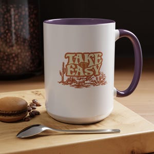 Puede incluir: Taza de cer&aacute;mica blanca con interior y asa morados. La taza presenta el texto "TAKE EASY" en una fuente retro con una ilustraci&oacute;n de paisaje des&eacute;rtico en tonos marrones y beige.