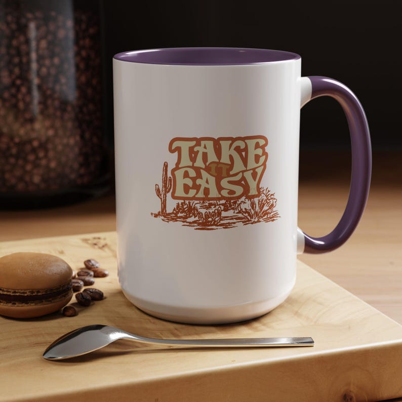 Puede incluir: Taza de cer&aacute;mica blanca con interior y asa morados. La taza presenta el texto "TAKE EASY" en una fuente retro con una ilustraci&oacute;n de paisaje des&eacute;rtico en tonos marrones y beige.