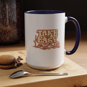 Puede incluir: Taza de cer&aacute;mica blanca con asa e interior azul marino. La taza presenta el texto "TAKE EASY" en una fuente retro con una ilustraci&oacute;n de paisaje des&eacute;rtico en tonos marr&oacute;n y beige.