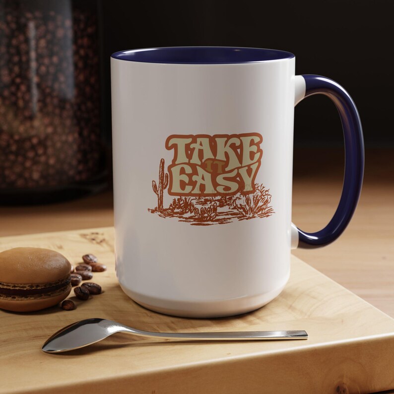 Puede incluir: Taza de cer&aacute;mica blanca con asa e interior azul marino. La taza presenta el texto "TAKE EASY" en una fuente retro con una ilustraci&oacute;n de paisaje des&eacute;rtico en tonos marr&oacute;n y beige.