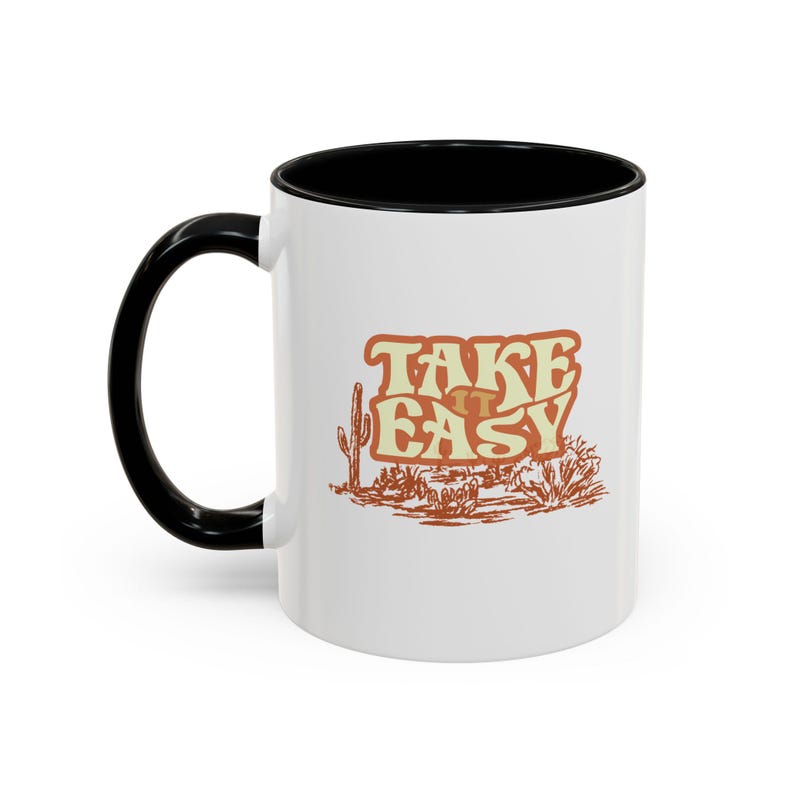 Puede incluir: Taza de cer&aacute;mica blanca con asa e interior negros. La taza presenta el texto "TAKE IT EASY" en un dise&ntilde;o naranja y marr&oacute;n con tem&aacute;tica des&eacute;rtica. Tambi&eacute;n se representa un cactus y un paisaje des&eacute;rtico.